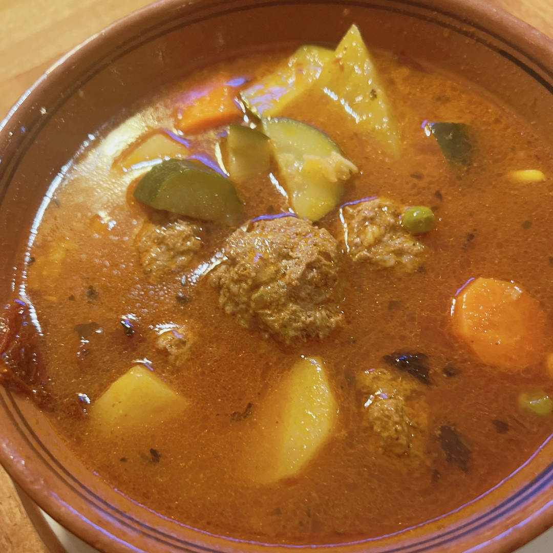 A bowl of albondigas soup, a traditional Mexican dish featuring meatballs in a rich, savory broth. The soup includes chunks of potatoes, zucchini, and carrots, with the vegetables and meatballs submerged in the flavorful red broth, creating a hearty and comforting meal.