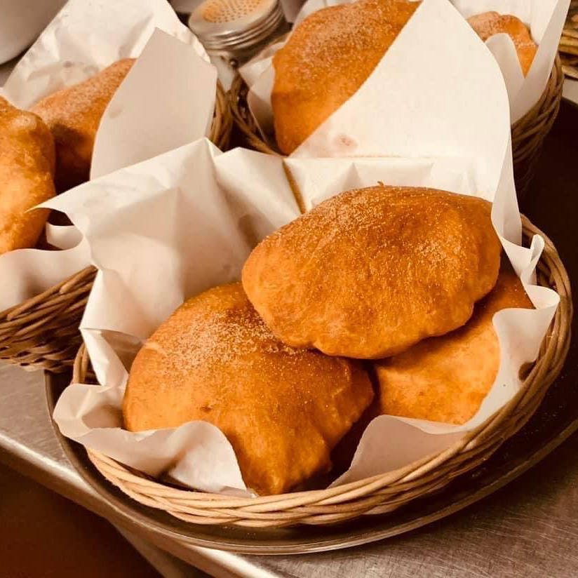 A close-up of baskets filled with golden-brown sopapillas, lightly dusted with sugar and lined with white parchment paper. The sopapillas are puffed and look freshly fried, sitting in woven baskets on a metal surface.