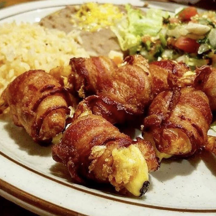 A plate of Camarones Rellenos, which are shrimp stuffed with cheese and wrapped in crispy bacon. The dish is served with sides of Mexican rice, refried beans topped with melted cheese, and a fresh salad of lettuce, tomatoes, and onions.