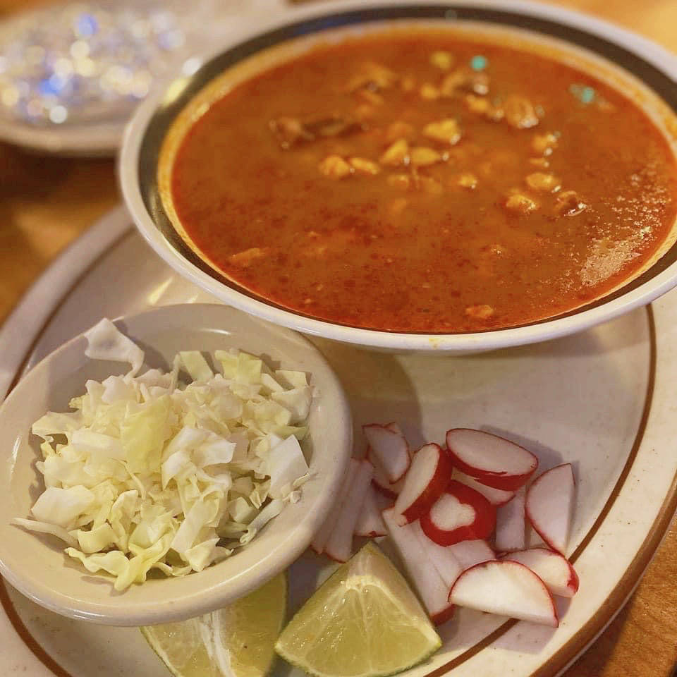 A bowl of pozole, a traditional Mexican soup made with a red, flavorful broth and hominy, served on a white plate. Accompanying the soup are side garnishes, including a small bowl of shredded cabbage, sliced radishes, and lime wedges, all placed on the plate next to the bowl.