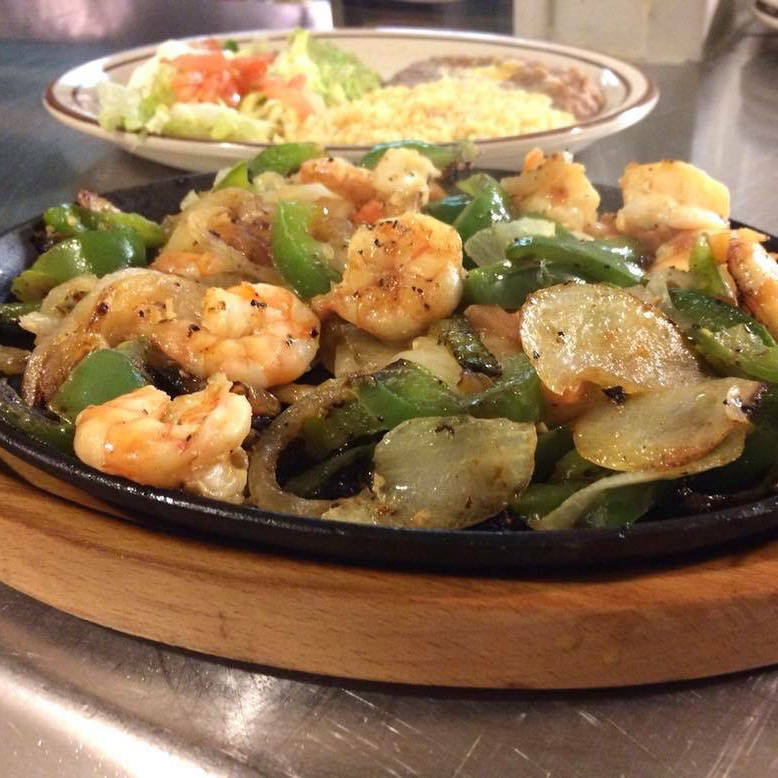 A sizzling platter of shrimp fajitas with sautéed onions and green bell peppers served on a wooden board. In the background, a plate with a side of rice, refried beans, and salad can be seen slightly out of focus.