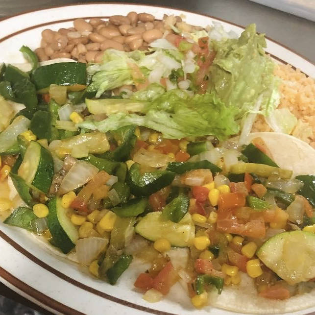 A plate of vegetarian tacos filled with a sautéed mix of zucchini, corn, bell peppers, onions, and tomatoes. The tacos are served with sides of Mexican rice, pinto beans, and a fresh salad with lettuce and diced tomatoes. The vibrant colors of the vegetables make the dish look fresh and appetizing