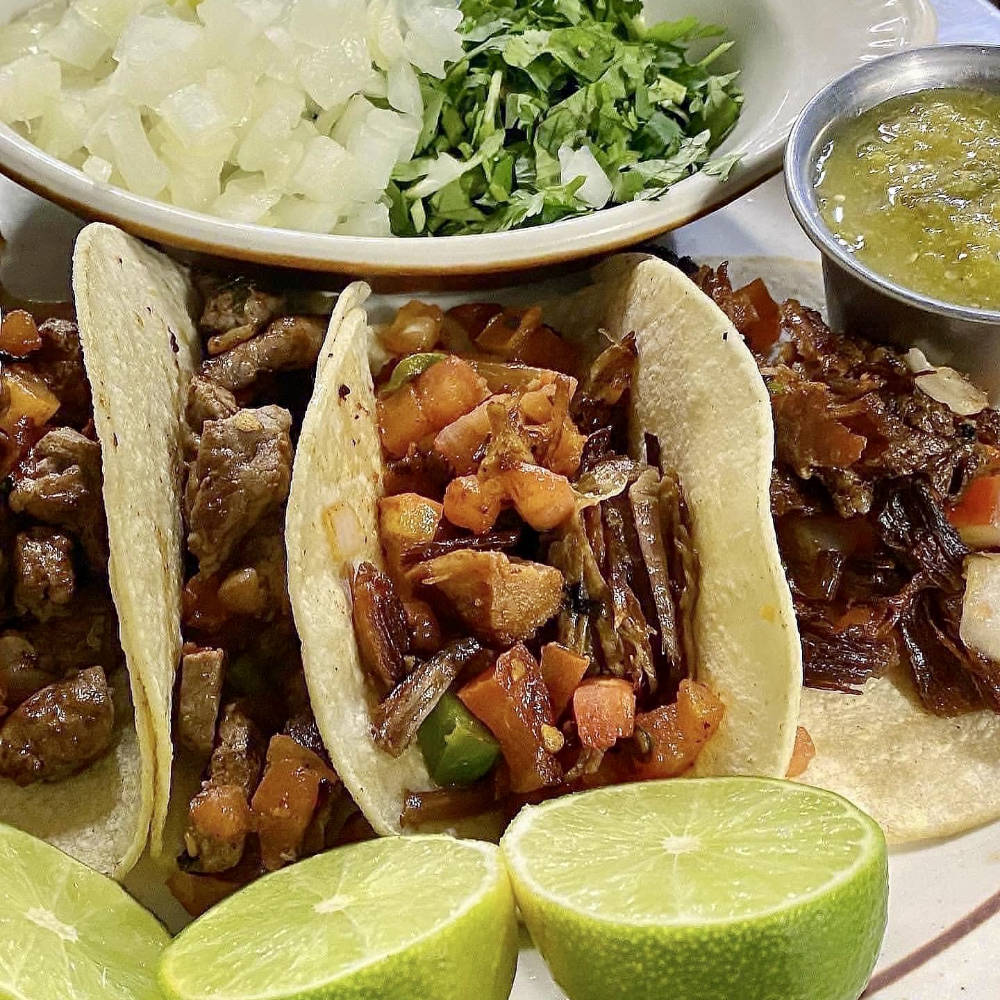 Three soft corn tacos filled with seasoned meat and a mix of sautéed vegetables, served on a plate with lime wedges. In the background, there is a small bowl with chopped onions and cilantro, and a metal cup with green salsa, adding the finishing touches to the taco dish.