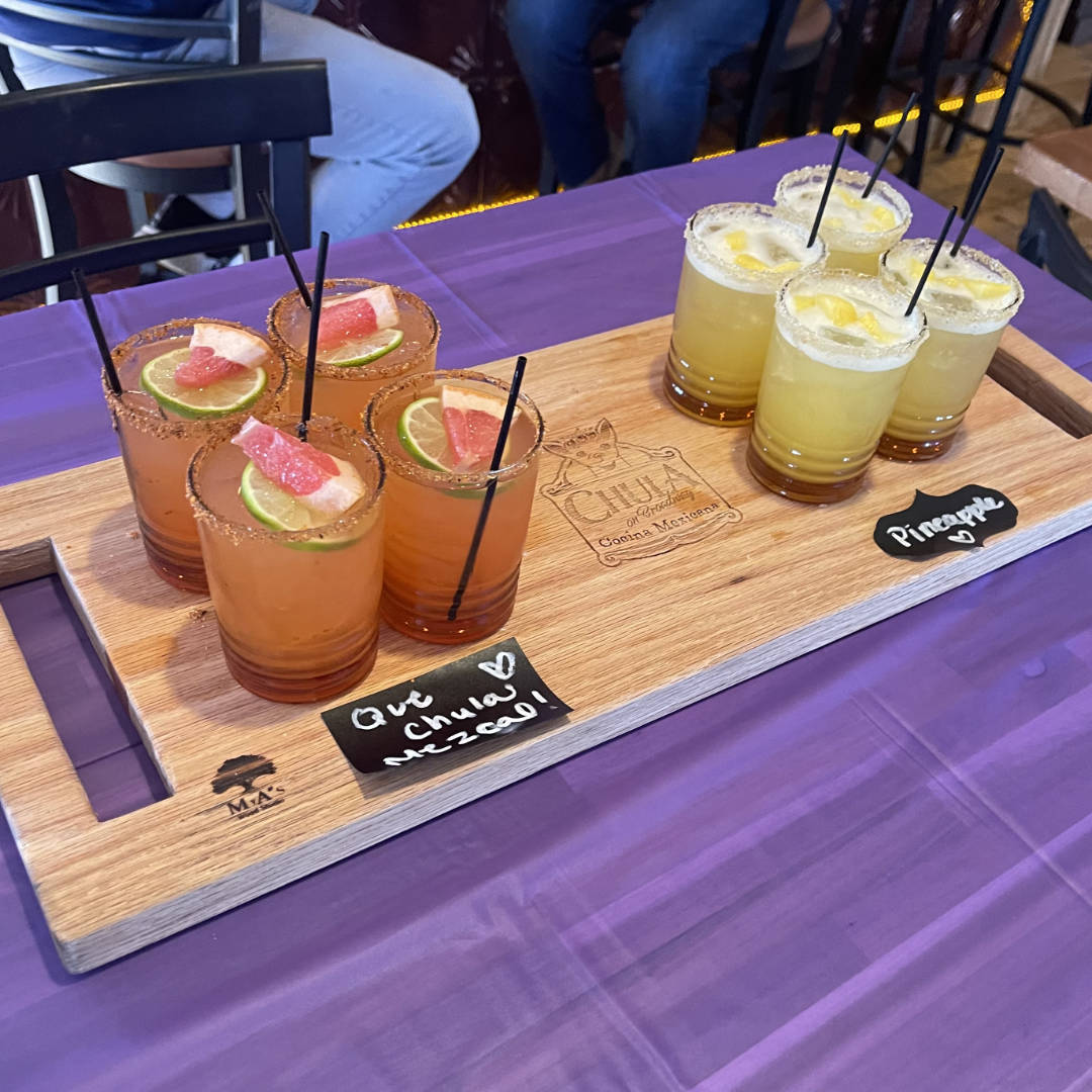 A wooden serving board featuring two sets of cocktails. On the left, four glasses filled with a reddish drink, garnished with lime slices and grapefruit wedges, and rimmed with a chili powder seasoning. On the right, four glasses with a yellow drink, garnished with lemon slices and a salt rim. The board has labels written in chalk, indicating 'Pineapple' for the yellow drinks and Qué Chula Mezcal for the reddish drinks. The board is set on a purple tablecloth, with a casual, festive atmosphere in the background.