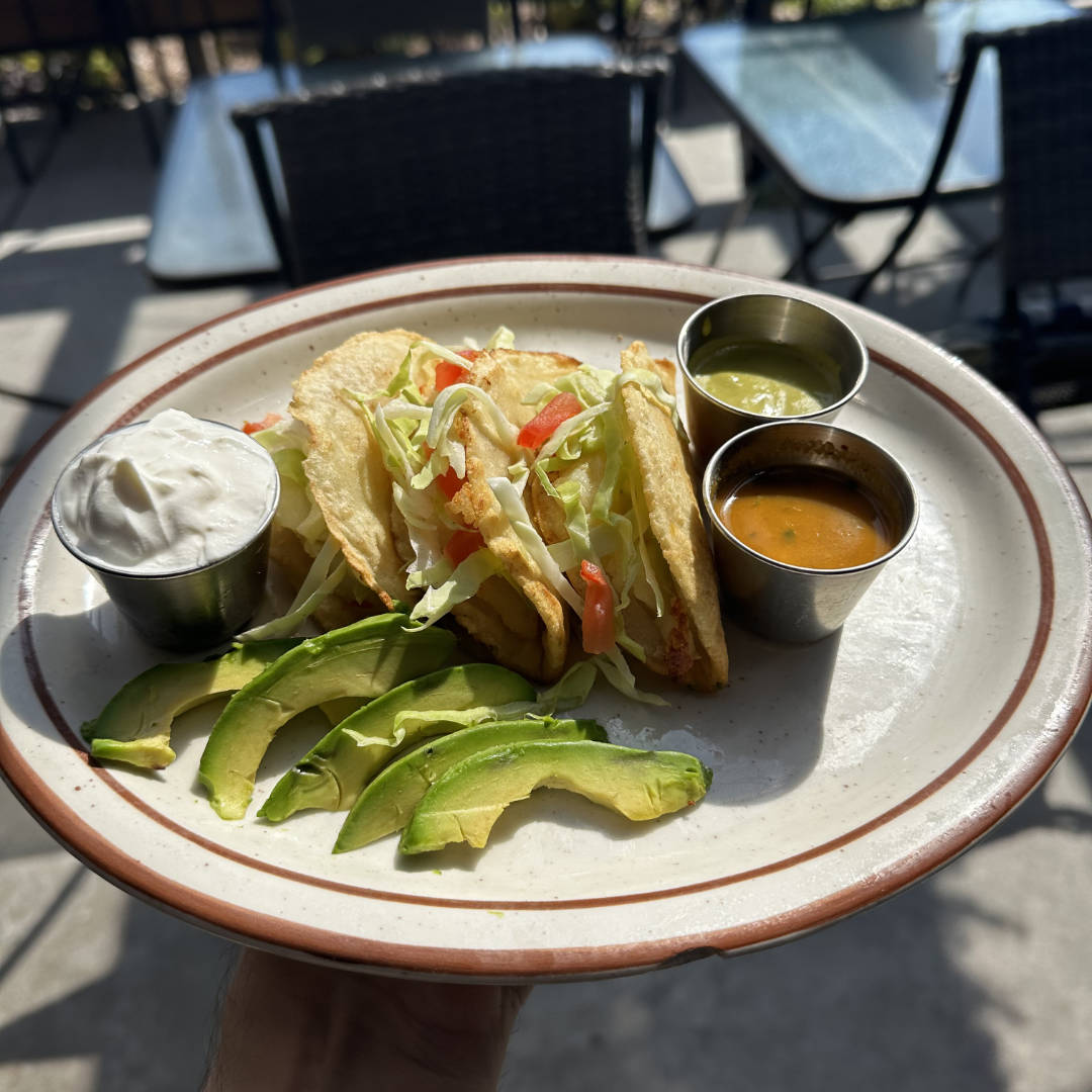 A plate of crispy tacos filled with shredded lettuce and diced tomatoes, served with slices of fresh avocado. The plate also includes small cups of sour cream, green salsa, and orange salsa as condiments. The dish is presented in an outdoor setting, with sunlight highlighting the fresh ingredients.