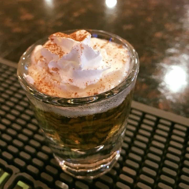 A shot glass filled with a layered cocktail, topped with a generous dollop of whipped cream and a sprinkle of cinnamon. The drink is placed on a bar mat with a dark, reflective surface in the background, giving it a festive and inviting appearance. This drink is called Chocolate Cake.