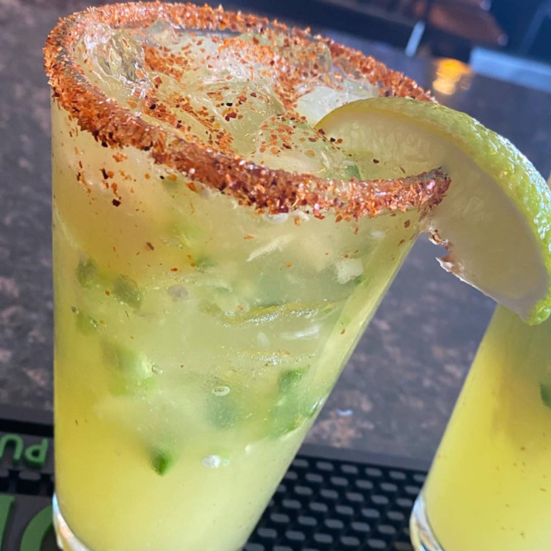 A close-up of a refreshing cocktail served in a glass with a chili-lime rim, garnished with a slice of lime. The drink is a vibrant yellow-green color, filled with ice and muddled ingredients, possibly mint or jalapeño, adding to its fresh appearance.