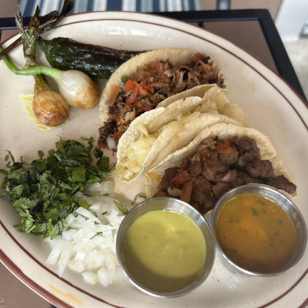 A close-up of three tacos filled with juicy, seasoned meat and sautéed vegetables, served in soft corn tortillas. The tacos are garnished with chopped onion and cilantro, and the plate is accompanied by halved lime wedges. In the background, a bowl with additional chopped onions and fresh cilantro is visible.