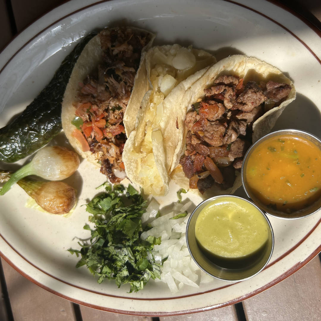 A platter of street tacos featuring small, soft corn tortillas filled with a variety of seasoned meats, topped with fresh onions, cilantro, and a sprinkle of lime juice. The tacos are accompanied by lime wedges and possibly a side of salsa, emphasizing the vibrant and fresh presentation typical of street-style tacos.