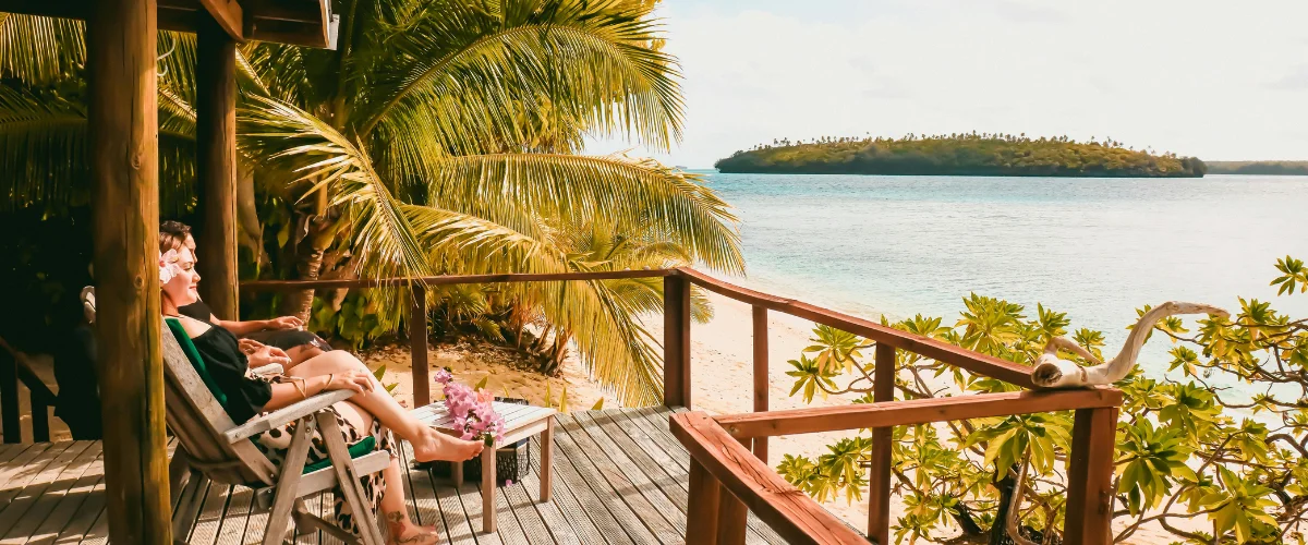 A couple relaxes on a wooden balcony overlooking a tropical beach with turquoise water and a distant island.