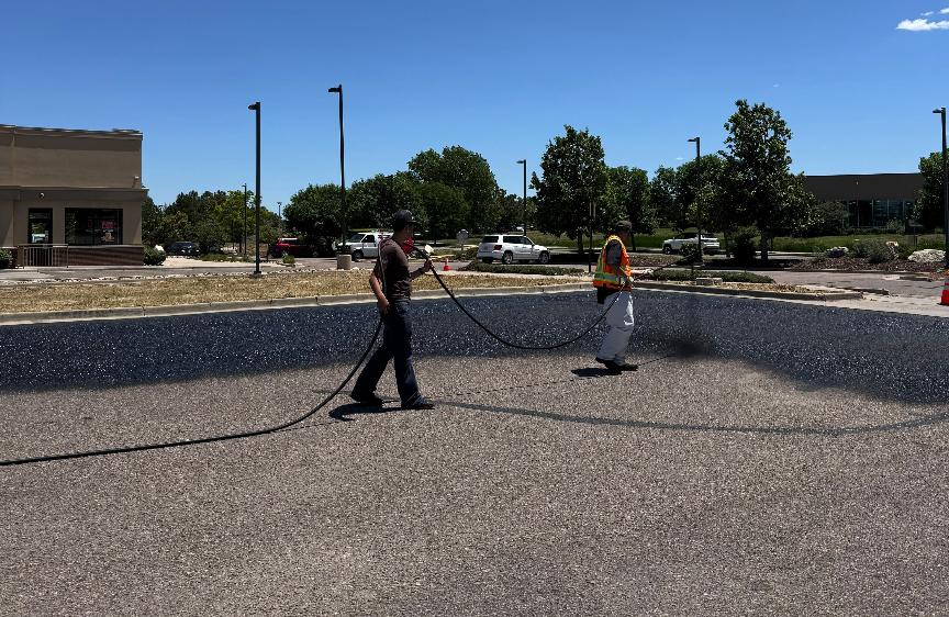 Two Elite crew members spraying sealcoat, with the parking lot half sealed and half untreated
