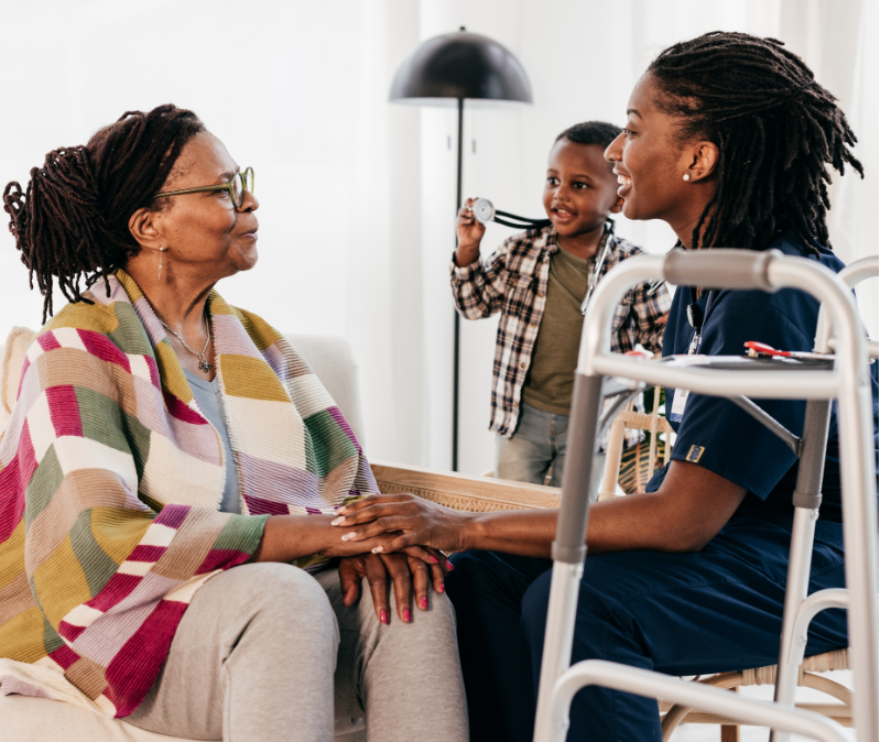 A nurse holding hands with an older woman at home while a child plays nearby