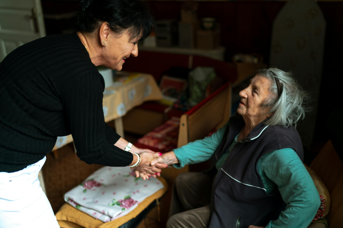 A nurse holding hands with an older woman at home while a child plays nearby