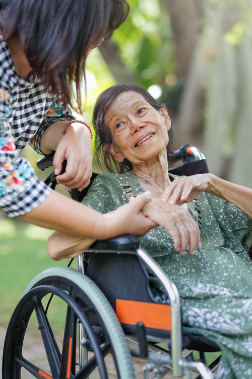A caregiver holding hands with an older woman beside her walker while a child plays nearby
