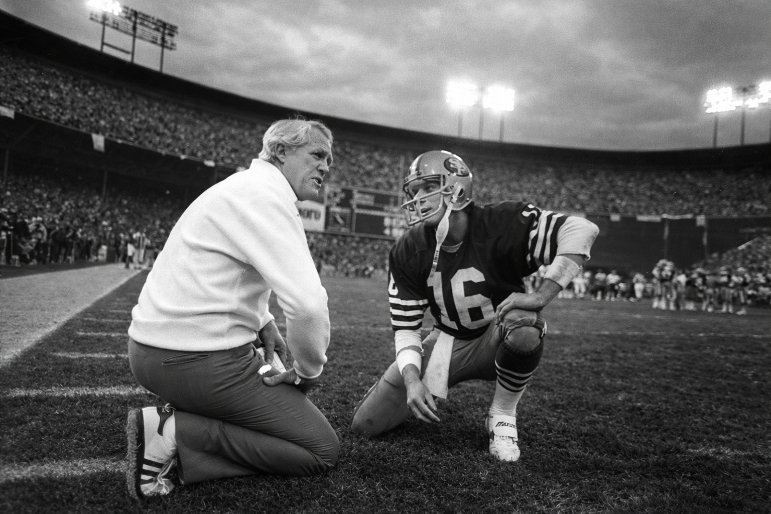 Black-and-white photo of an American football coach talking with a quarterback wearing number 16 on the sideline during a game, with a packed stadium and bright stadium lights in the background.