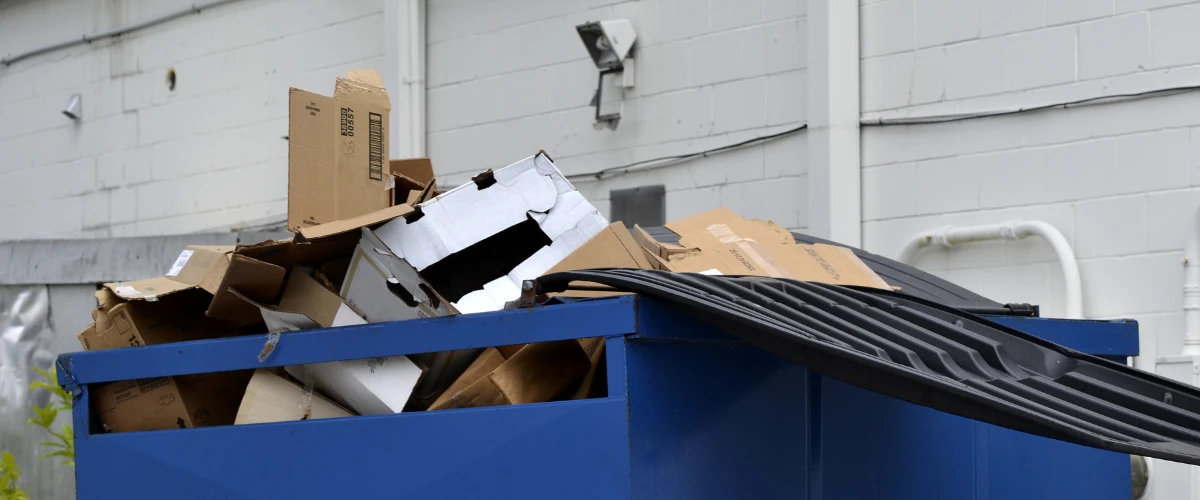 Overflowing blue dumpster filled with flattened cardboard boxes behind a commercial building.