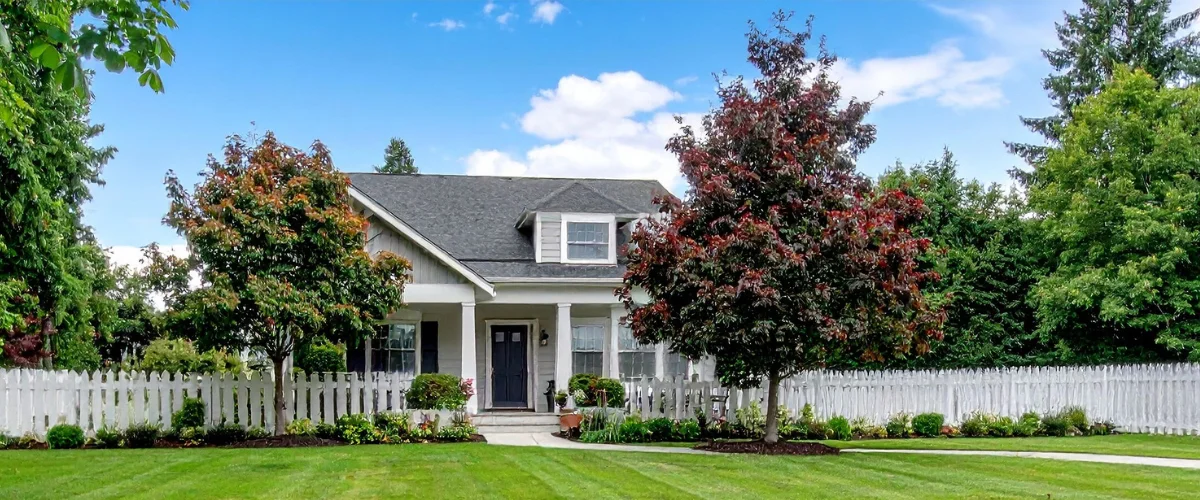 A classic gray two-story suburban house with a dark roof and a front porch, framed by a white picket fence and lush green lawn.