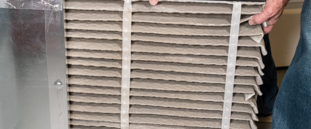 A person's hands installing or removing a dirty pleated air filter from an HVAC unit, with silver ductwork visible.