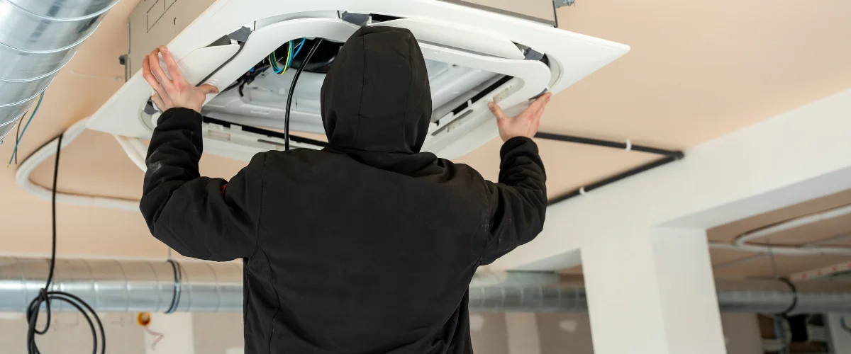 A person in a black hoodie installing a ceiling-mounted HVAC unit, showing hands on the unit and exposed wires.