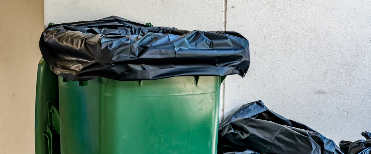 A close-up shot of a green garbage can with a black plastic liner overflowing from the top.