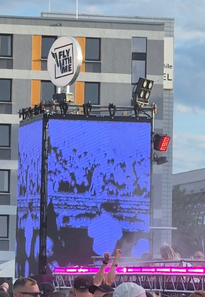 Bühne beim Fly-with-me-Festival am Abend: ein rundes Schild mit der Aufschrift Fly with me über einer blau leuchtenden LED-Wand, davor die Silhouetten der Besucher.