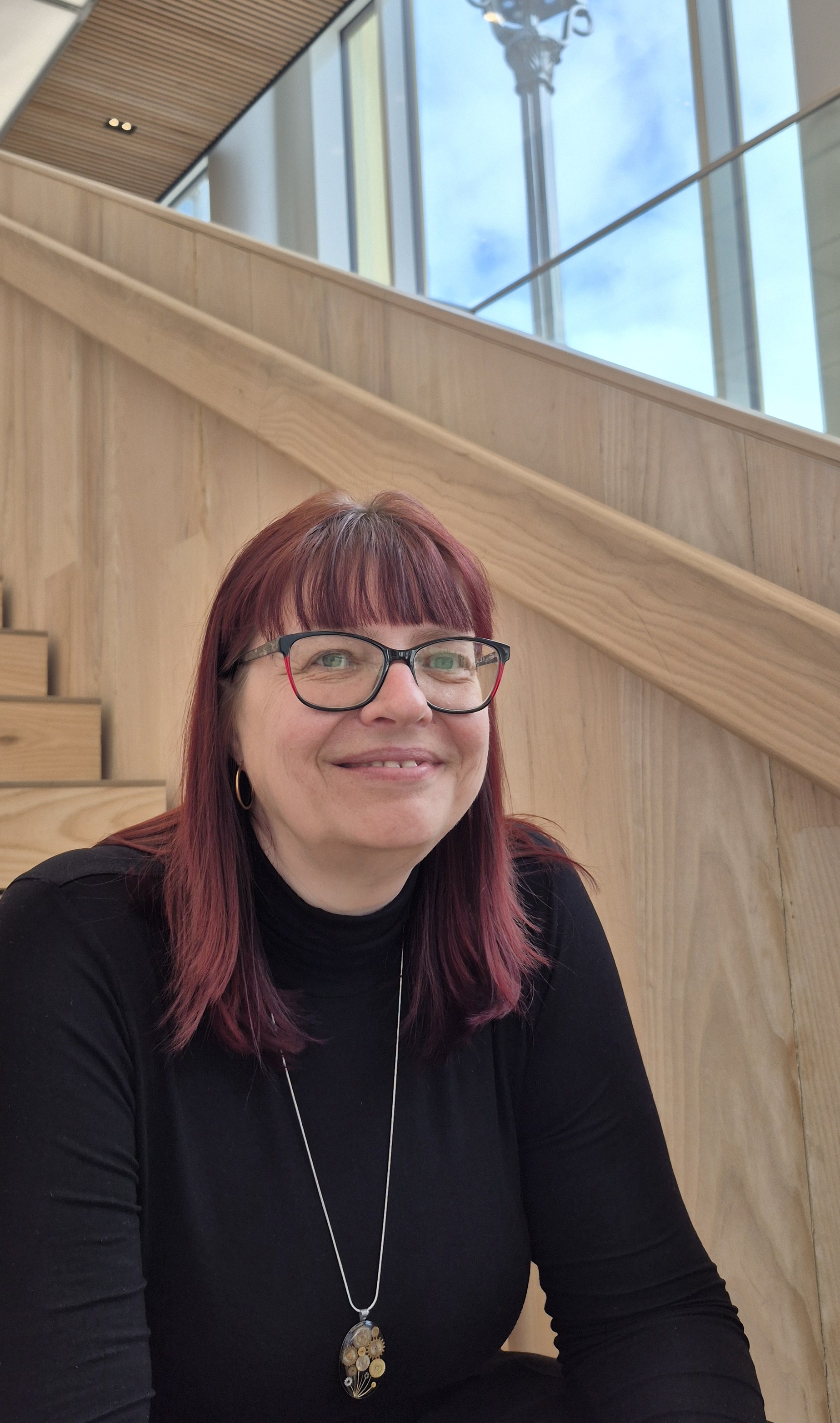 Marion Schumann sitzt lächelnd auf einer hellen Holztreppe, hinter ihr eine große Fensterfront mit blauem Himmel.
