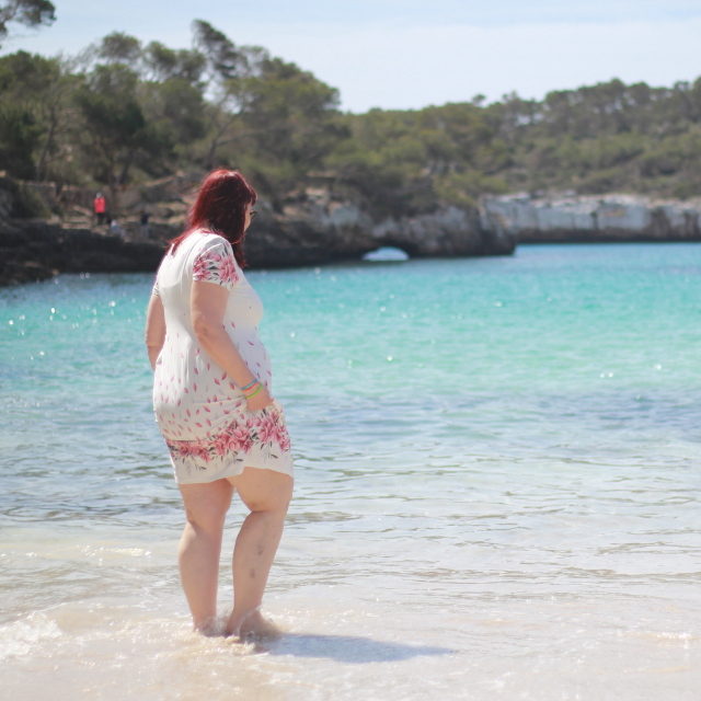 Frau mit roten Haaren in einem hellen, geblümten Sommerkleid steht knöcheltief im türkisfarbenen Wasser einer Bucht und blickt ruhig aufs Meer hinaus.