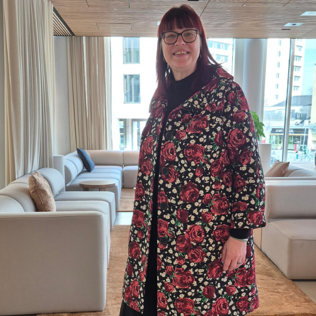 Marion steht lächelnd in einer hellen Lounge mit großen Fensterfronten und beigen Sofas, trägt einen langen Mantel mit rotem Rosenmuster und schaut direkt in die Kamera.