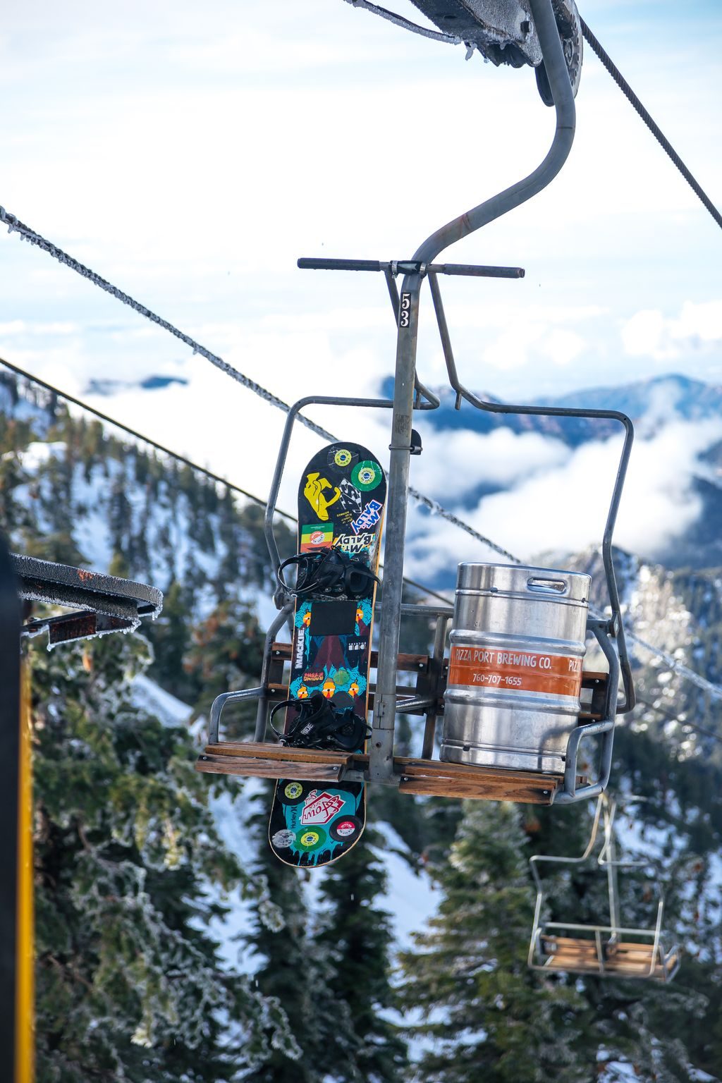 A snowboard and a beer keg riding a chair above snowy pines and clouds