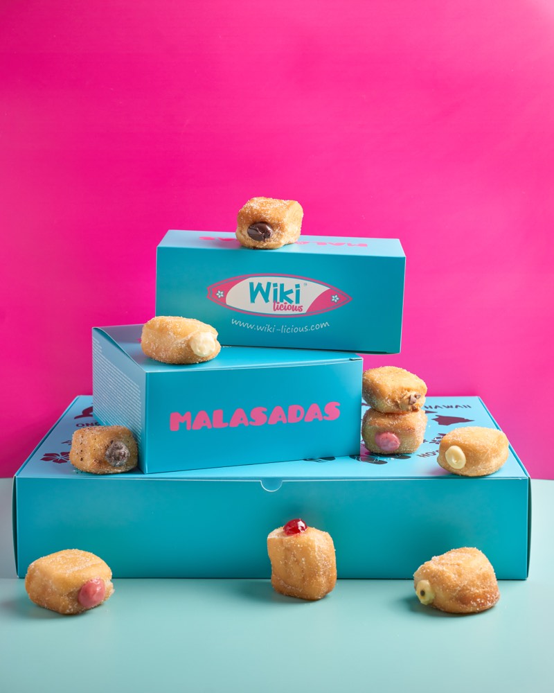Three stacked teal Wiki-Licious boxes with sugar-dusted malasadas perched on top, shot against a hot-pink backdrop