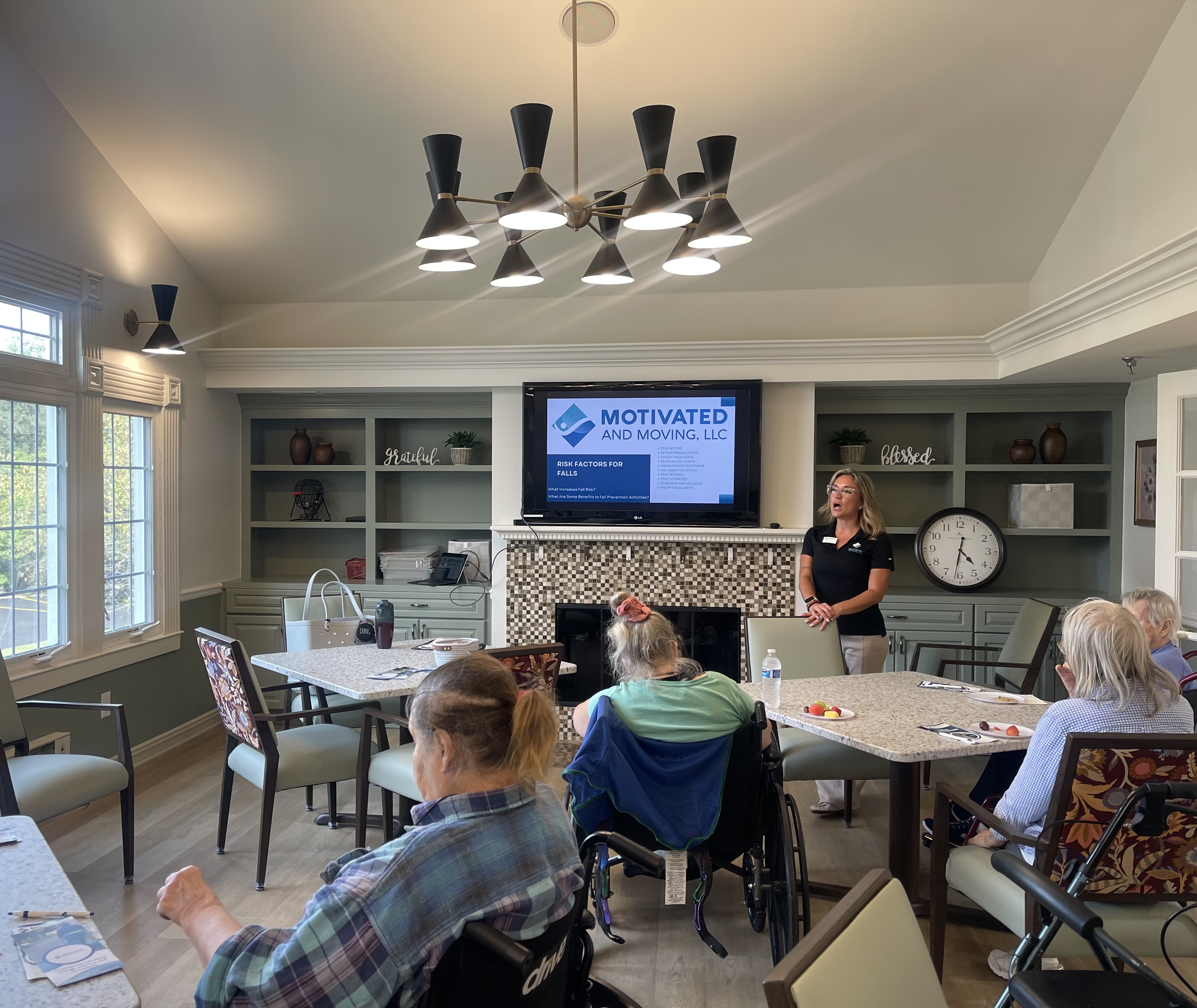 Jasmine leading a live wellness session in a community room while seated residents follow along, viewed from behind.