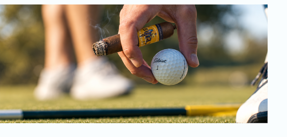 A golfer holding a BULLY Maduro cigar picks up a golf ball beside a putter on the green.