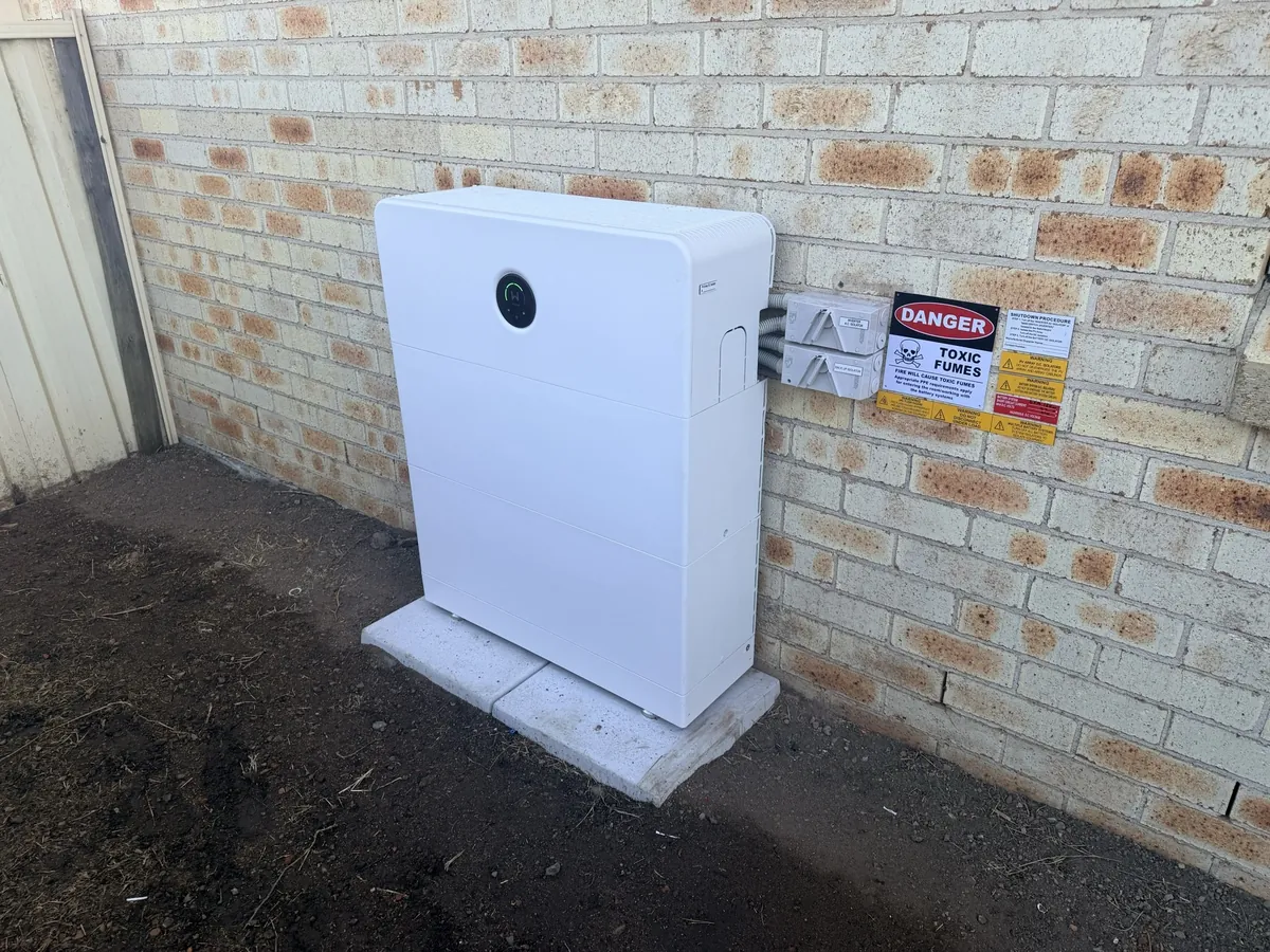 Home battery installed by Dunamis Power on a brick wall, with isolators and safety signs beside it