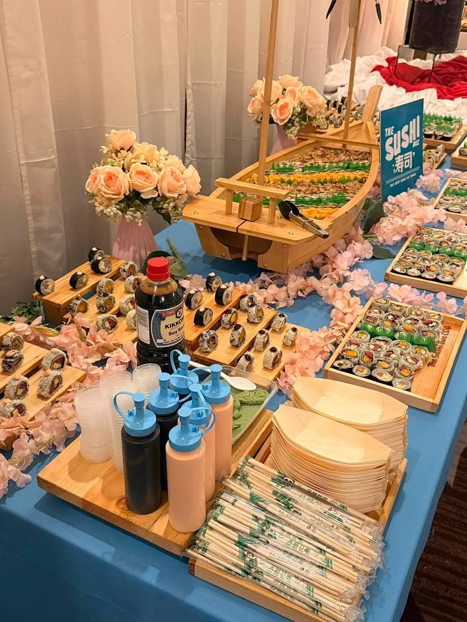 Sushi boat catering spread with maki trays, condiments, and floral decor