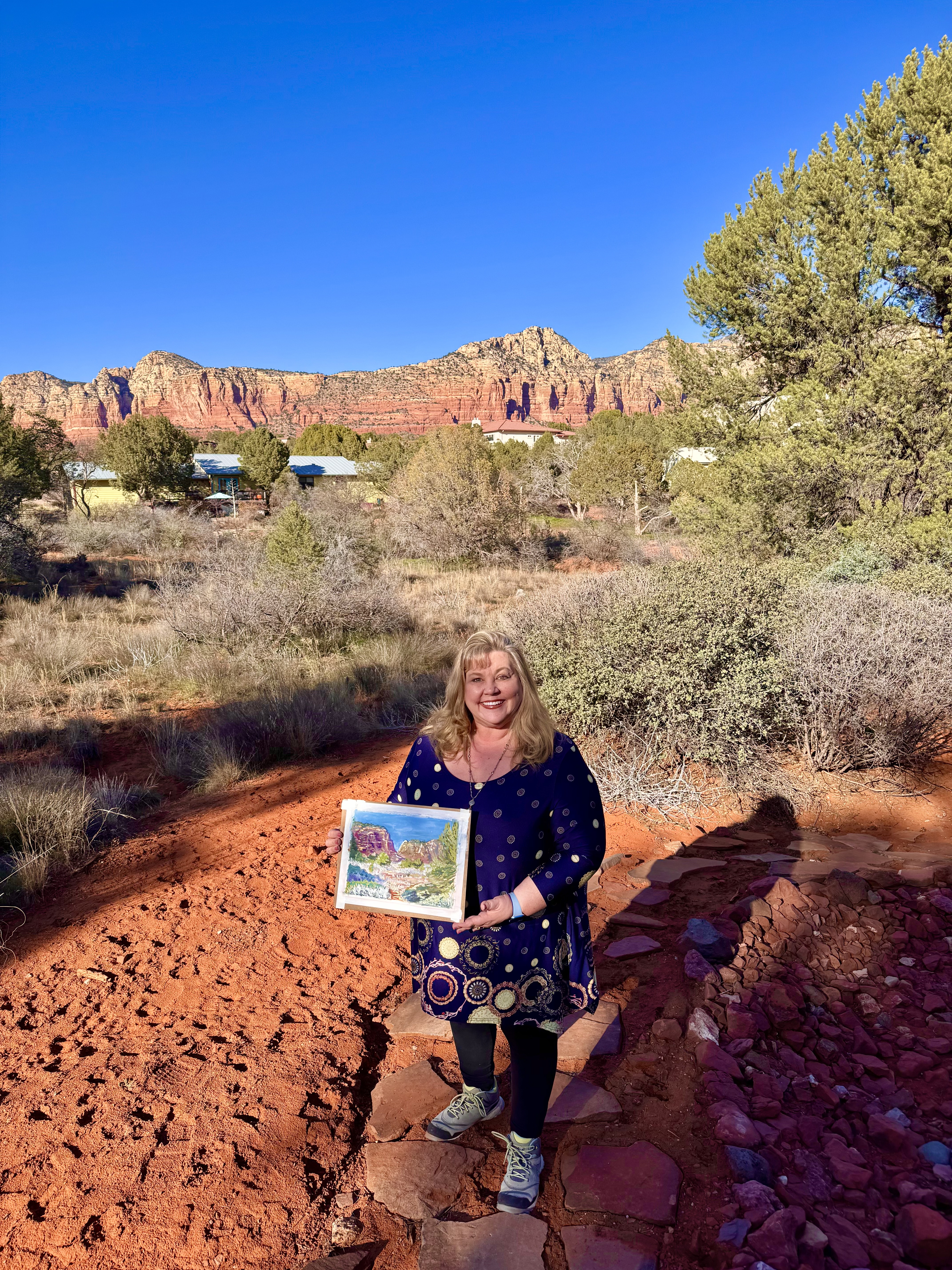Artist Elizabeth Van Tassel holding artwork painted in plein air in Sedona with red rock formations around.