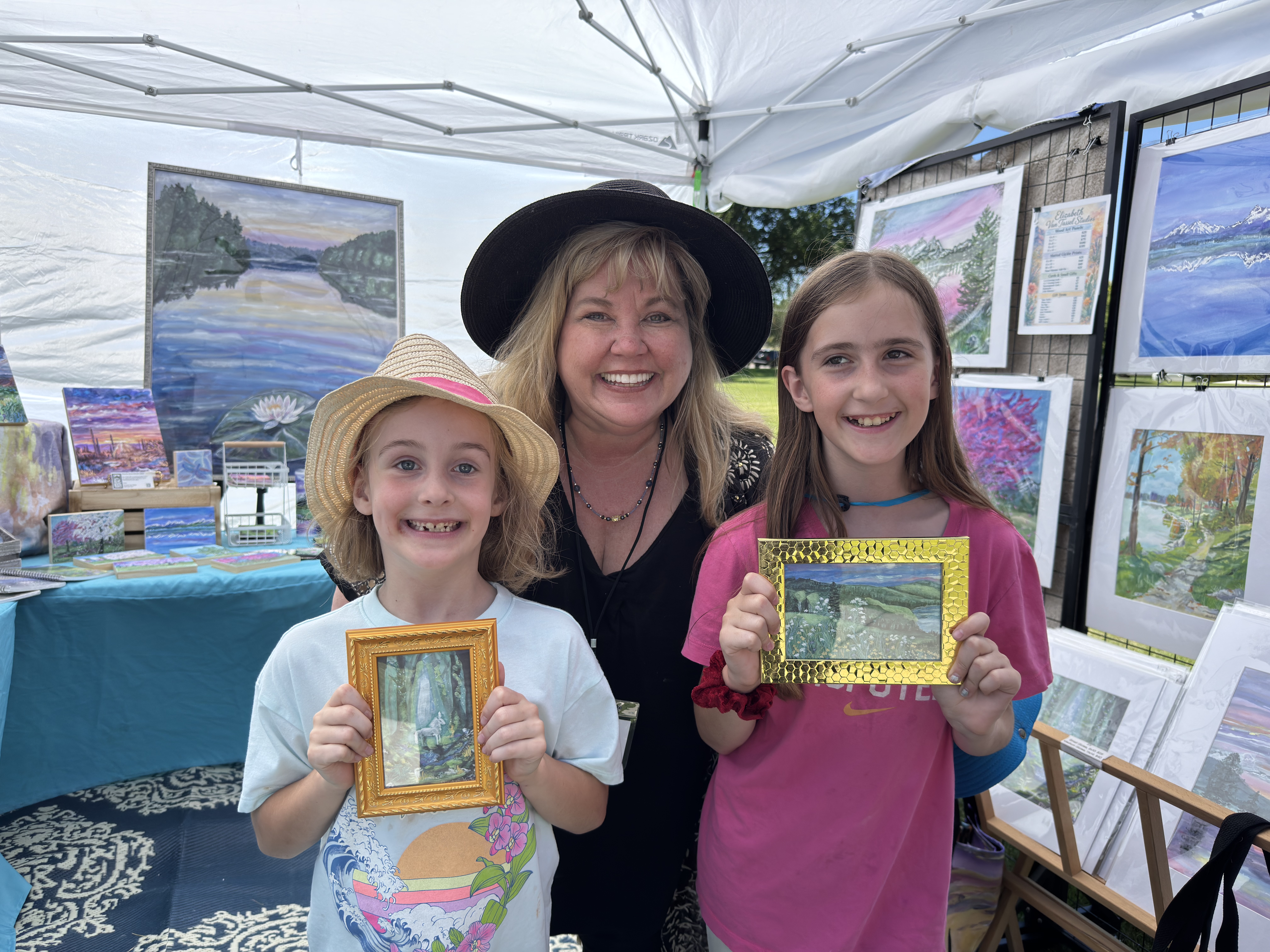 Artist Elizabeth Van Tassel smiling with two young collectors in her outdoor art booth as they hold small framed Shimmers of Light artworks, a sweet moment of children connecting with original art.