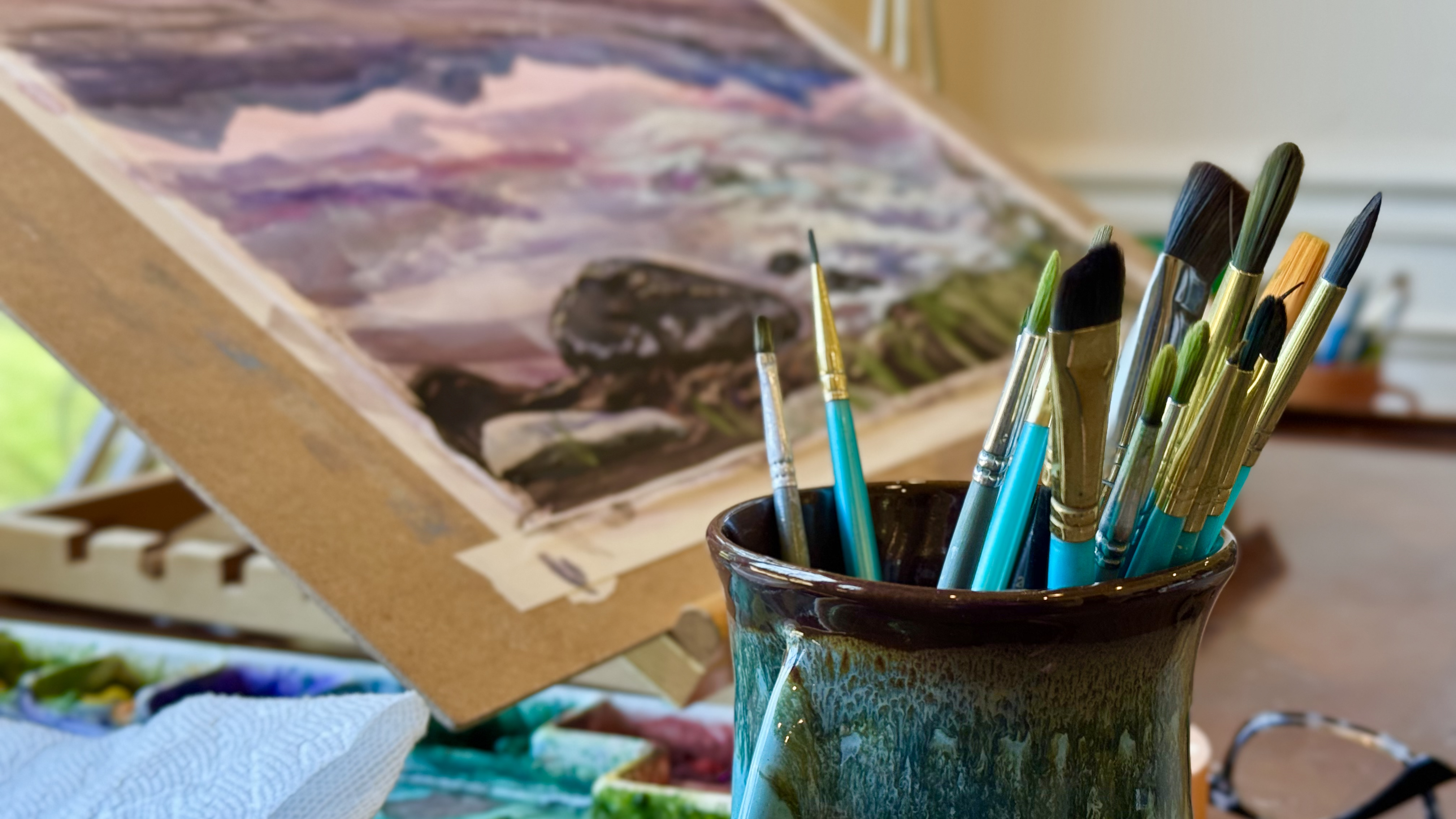 Paintbrushes in a ceramic cup beside a watercolor landscape painting in progress, showing the working studio process of artist Elizabeth Van Tassel.