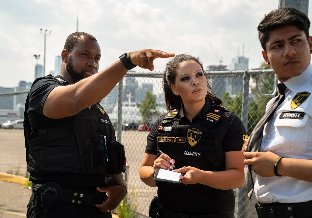 Security officers checking a chain-link fence line, one taking notes