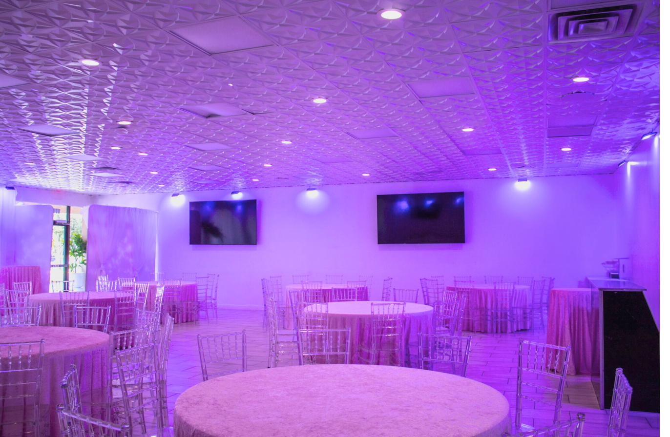 The full ballroom under purple uplighting, round tables set with clear chiavari chairs
