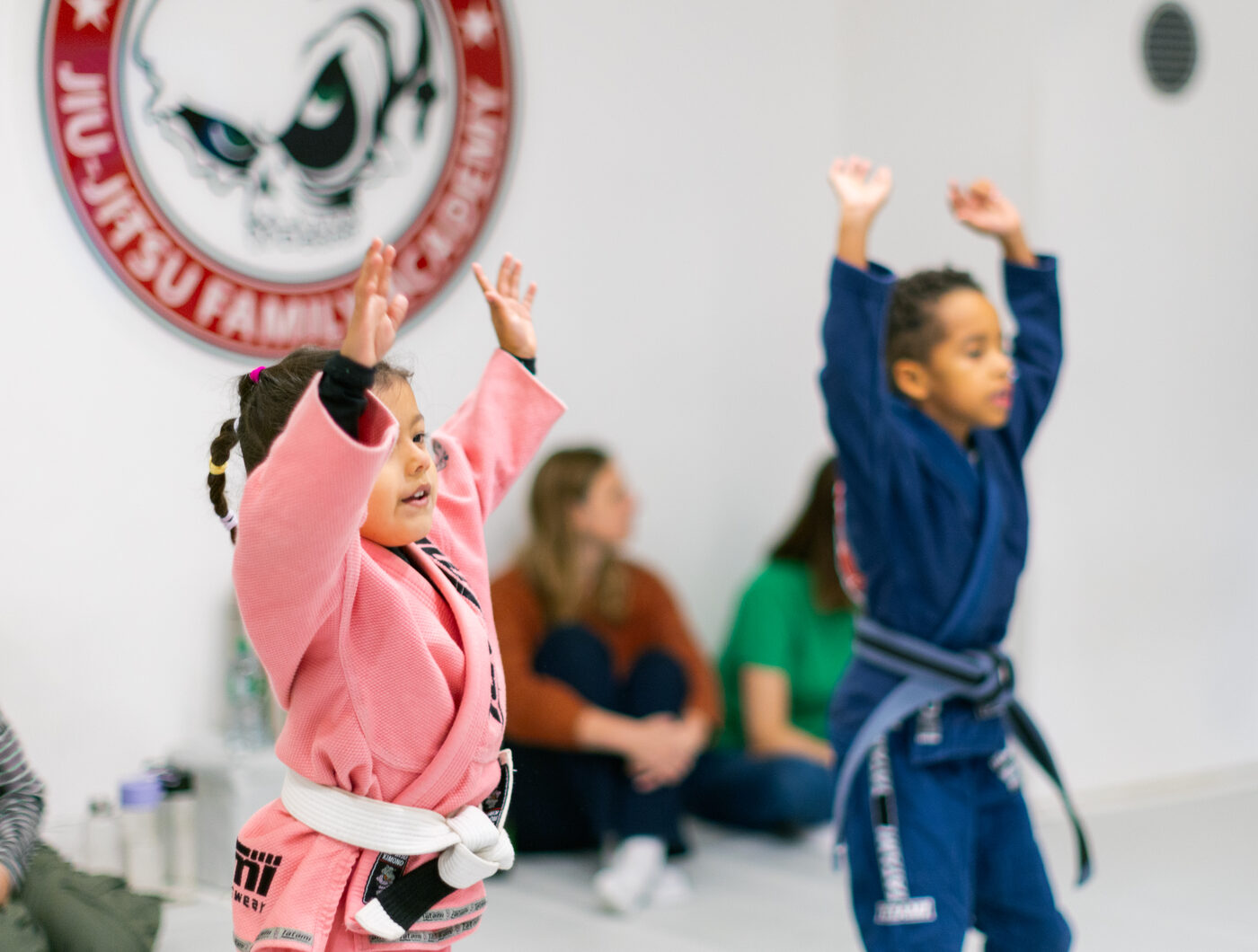 Zwei Kinder im CJJF-Trainingsraum in Düsseldorf strecken lachend die Arme in die Höhe, im Hintergrund sitzen begleitende Elternteile