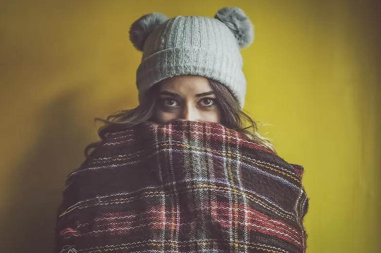 Woman in a grey pom-pom beanie with a tartan scarf pulled up to her nose against a yellow wall