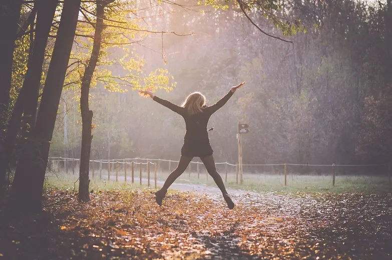 Woman leaping with arms outstretched on a leaf-covered path in a sunlit autumn forest
