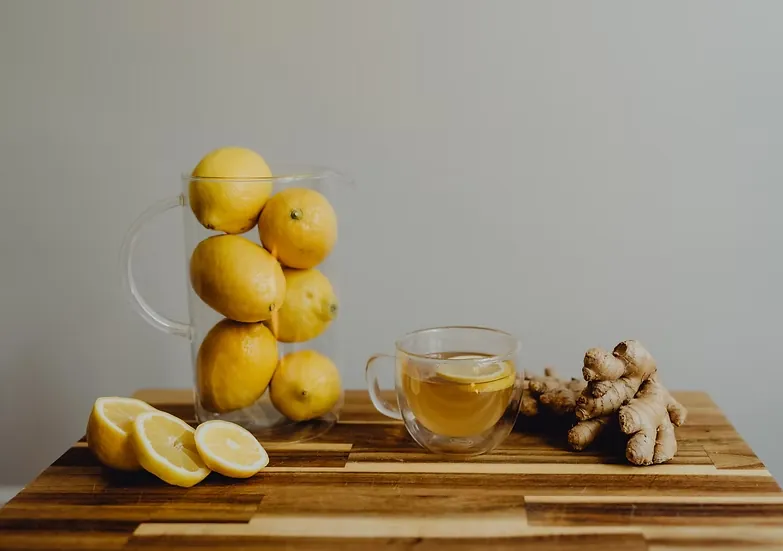Glass cup of lemon and ginger tea beside fresh ginger root, lemon slices and a jug of whole lemons