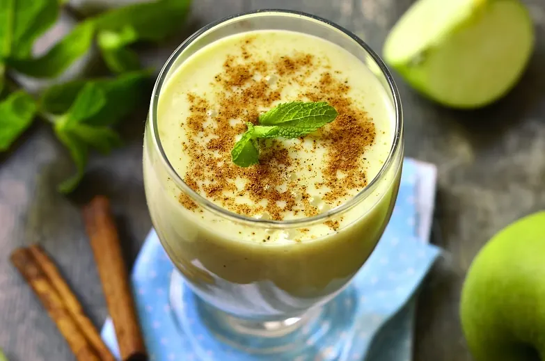 Green apple smoothie topped with cinnamon and a mint leaf, beside cinnamon sticks and apples