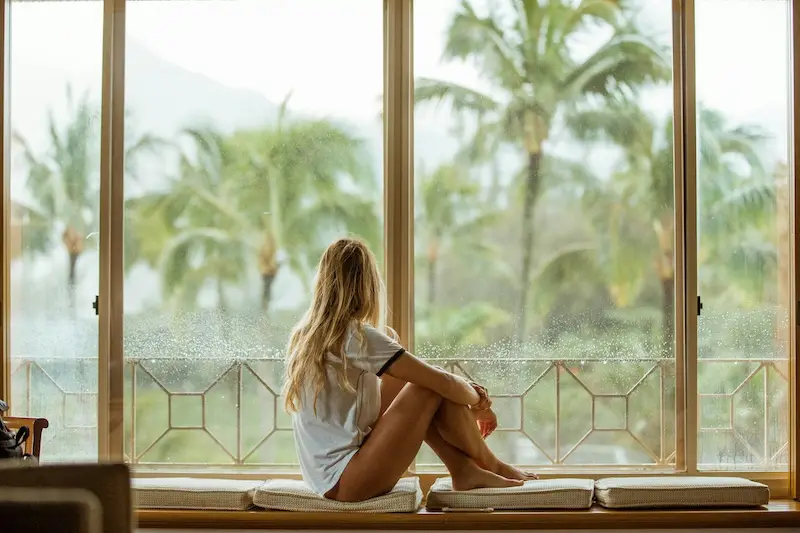 Woman sitting by a large window looking out at palm trees