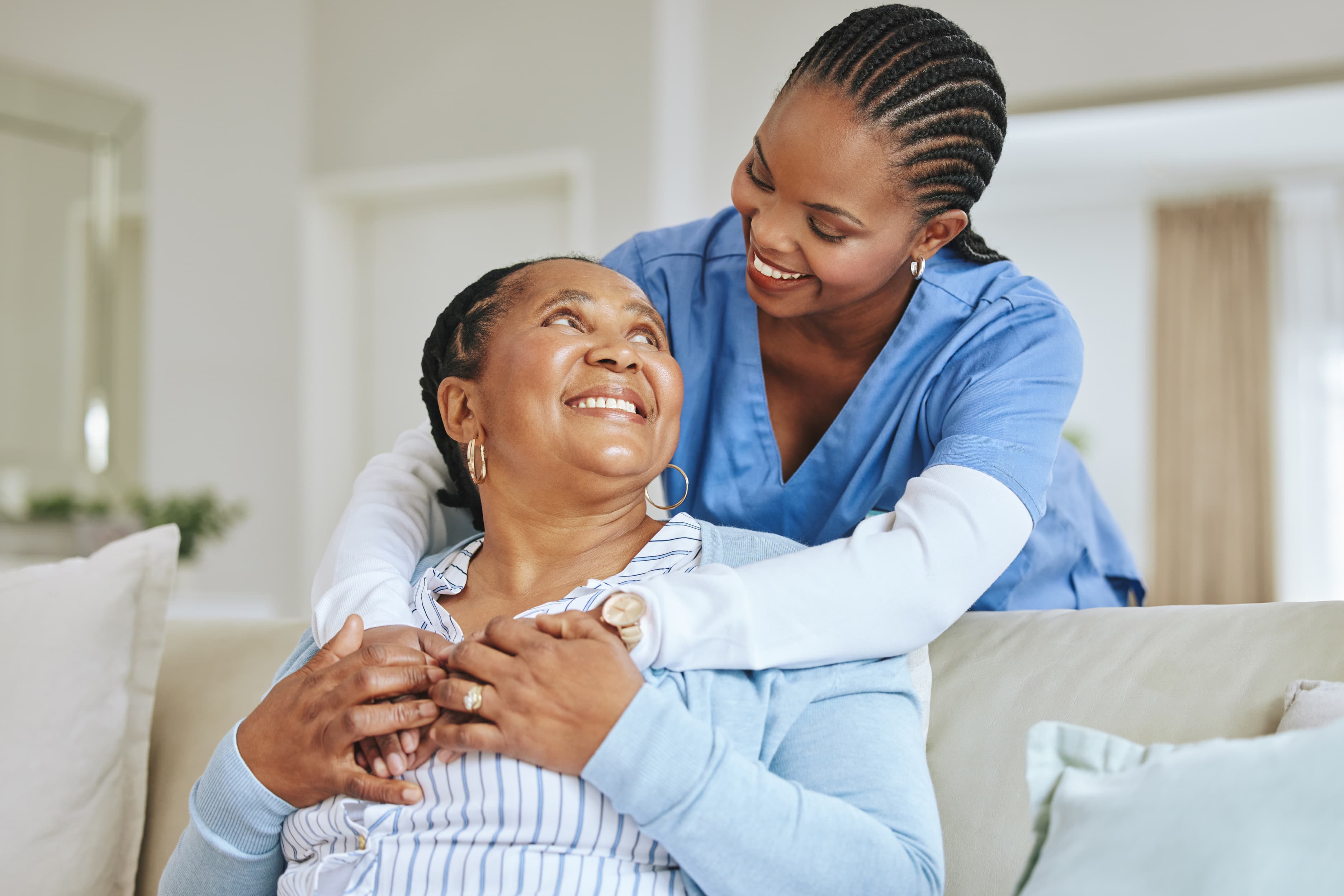Healthcare professional in blue scrubs warmly embracing an elderly woman on a couch in a bright living room, reflecting compassion and supportive care.