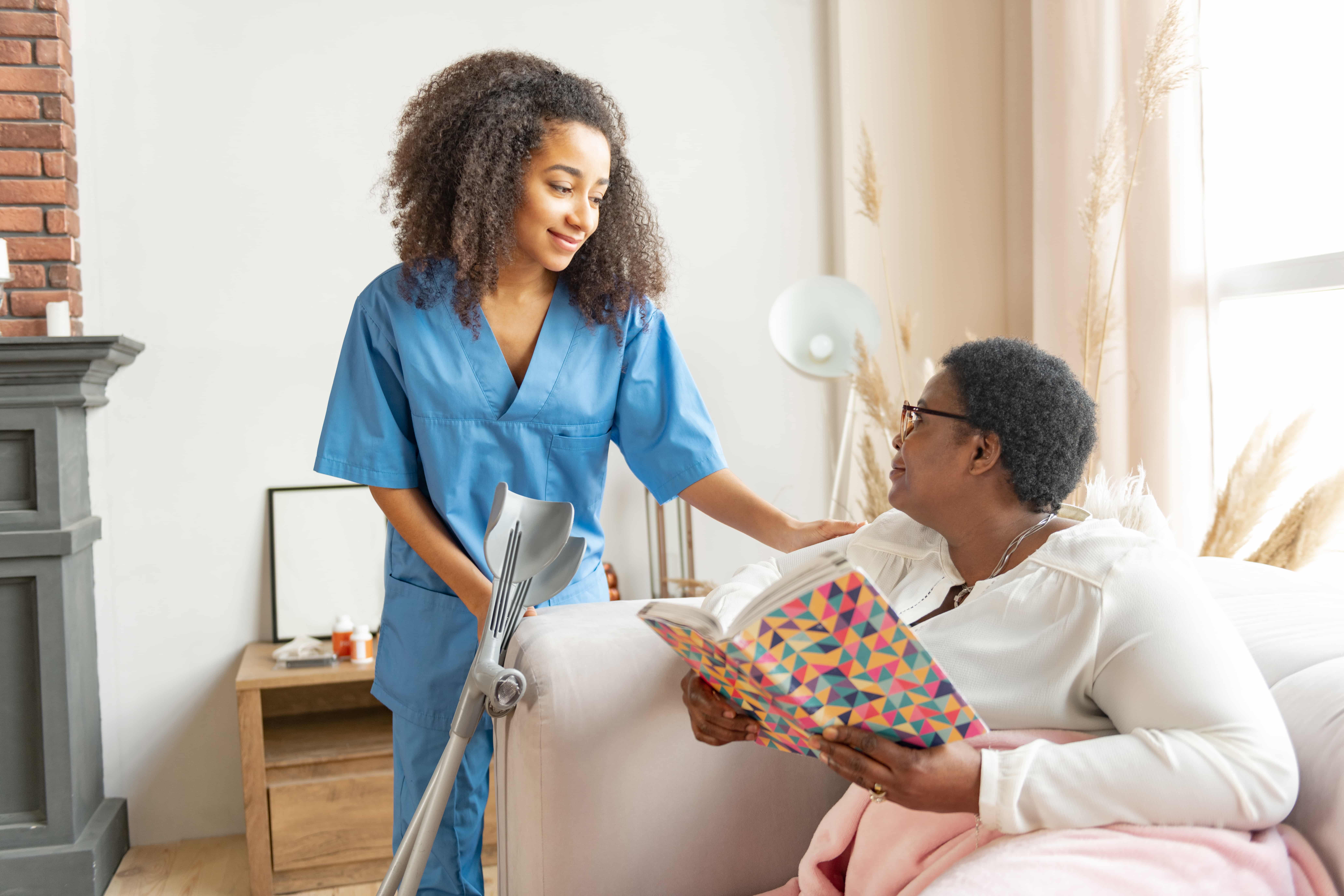 Caregiver in blue scrubs gently placing a hand on the shoulder of an elderly man seated with a patterned notebook and pink blanket, with crutches nearby in a warm home care environment. Caregiver in blue scrubs gently placing a hand on the shoulder of an elderly man seated with a patterned notebook and pink blanket, with crutches nearby in a warm home care environment.
