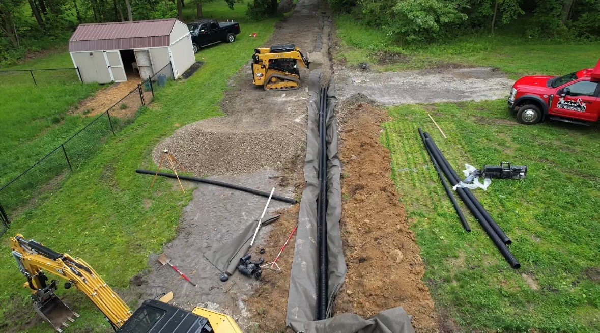 Excavator and skid steer installing drainage trench with protective fabric and gravel base