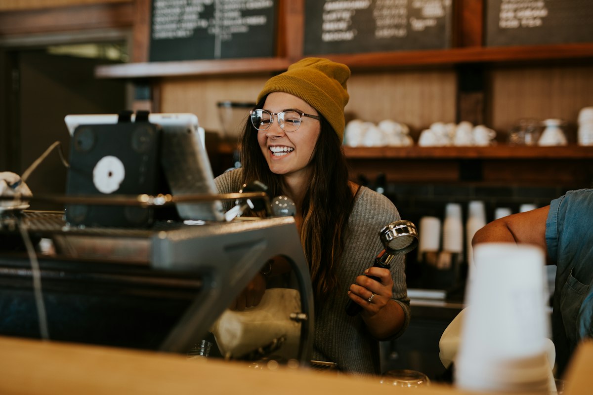 A smiling woman in a mustard beanie holding a portafilter in a coffee shop
