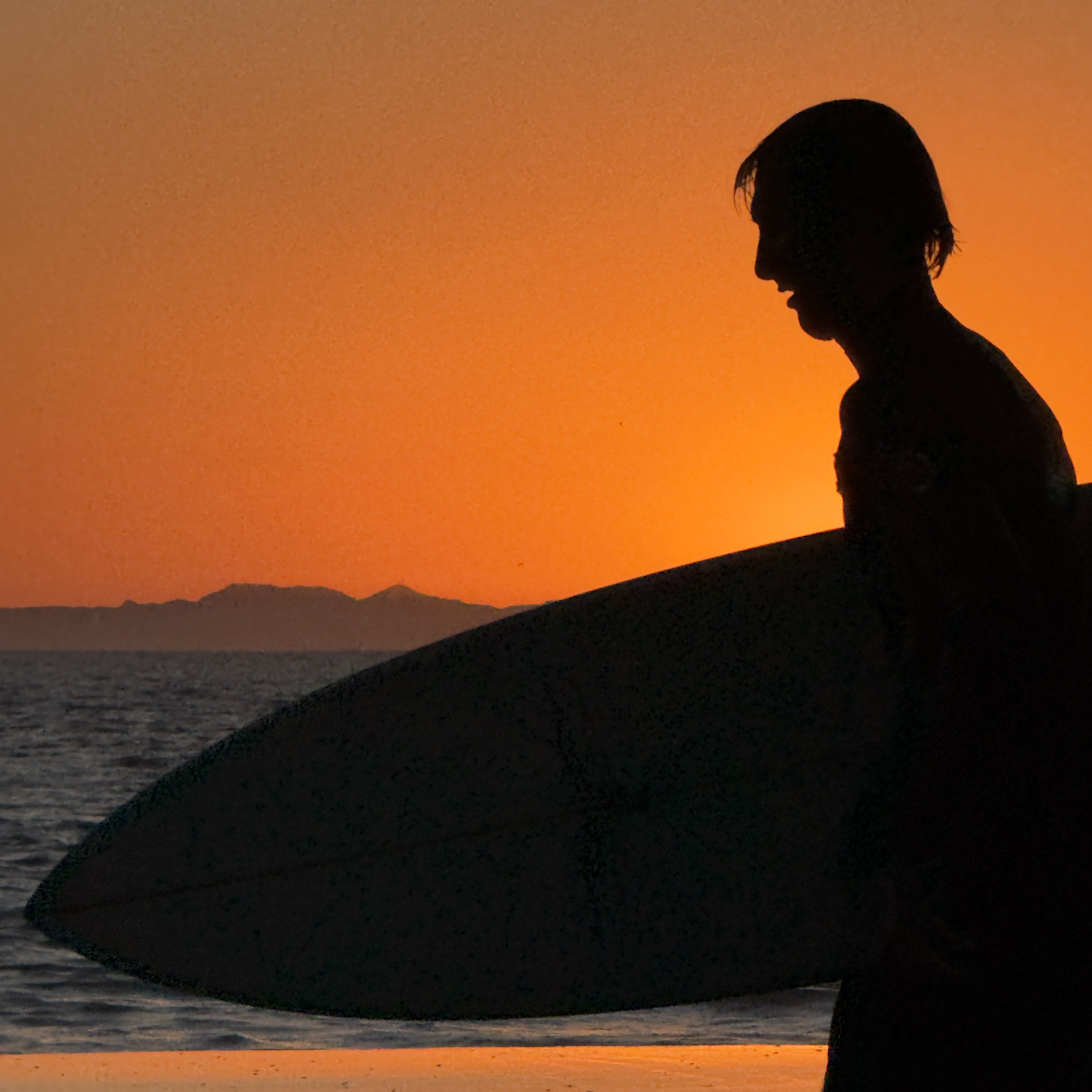 Silhouette of a surfer holding a surfboard at sunset, with the ocean and a distant island under an orange sky.